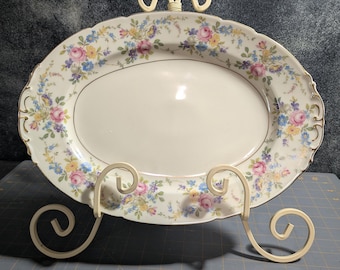 May include: An oval porcelain serving platter featuring a cream-colored center and a decorative border adorned with pink roses, yellow blossoms, and blue forget-me-nots, finished with a scalloped edge and gold trim, displayed on a metal stand.
