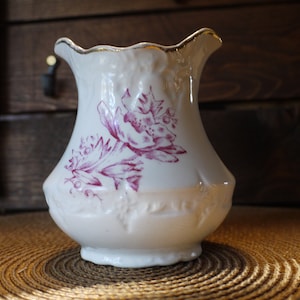 May include: A white porcelain creamer with a scalloped rim and a pink floral design. The creamer has a gold trim around the rim.