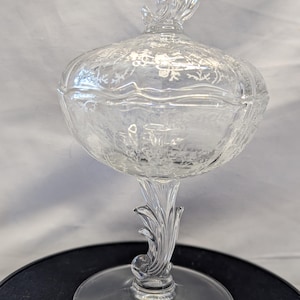 May include: A clear glass candy dish with a footed base and a lid. The dish is decorated with a delicate etched floral pattern.