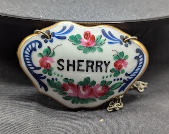 May include: A white porcelain bottle stopper with a gold trim and blue and green floral designs. The stopper has a chain attached and the word "SHERRY" written in black lettering.