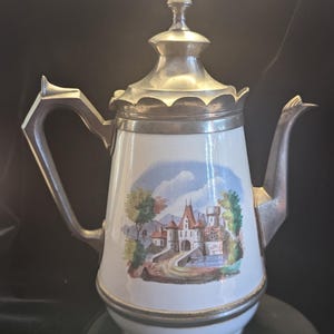 May include: A white porcelain teapot with a silver lid and handle. The teapot features a painted scene of a castle with a bridge and trees.