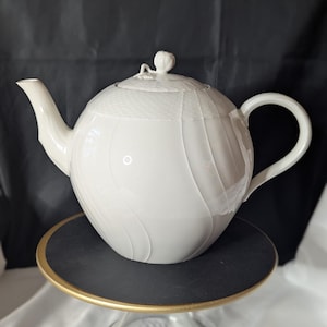 May include: A white ceramic teapot with a rounded body and a textured lid. The teapot has a curved spout and handle. It is displayed on a black and gold cake stand with a clear glass base. The background is black.