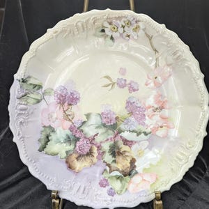 May include: A vintage porcelain plate with a delicate floral design featuring pink and purple flowers, green leaves, and purple berries. The plate has a scalloped edge and a white background with a light purple wash.