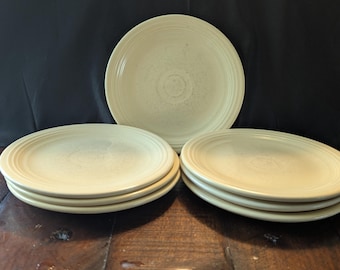 May include: A collection of cream-colored ceramic plates. The plates have a simple, elegant design with concentric rings around the edges. The plates are stacked in sets of four and five, with one plate standing alone in the background.
