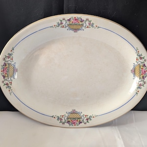 May include: Oval serving platter with a cream-colored surface, adorned with floral basket designs at each end and a blue line border. The rim is trimmed with gold. The floral baskets feature pink, yellow, and red flowers.