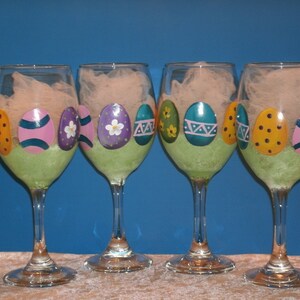 May include: Four clear glass wine glasses with green painted bases and painted Easter egg designs. Each glass features three eggs with different colors and patterns.