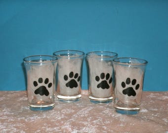 Puede incluir: Cuatro vasos de chupito transparentes con un diseño de huella de pata negra. Los vasos están llenos de material blanco y están colocados sobre un fondo rosa y azul. Un artículo divertido para los amantes de las mascotas.