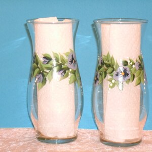 May include: Two clear glass vases with a hand-painted design of green leaves and purple flowers. The vases are approximately 10 inches tall.