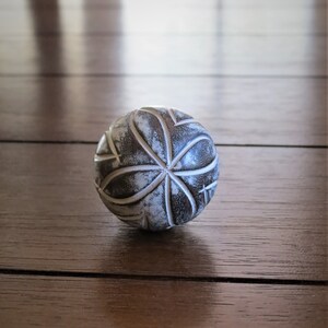 Op de afbeelding: Een witte en grijze decoratieve bal met een gegraveerd geometrisch patroon. De bal ligt op een houten oppervlak.