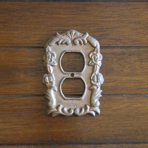 May include: A decorative cast iron light switch cover with a floral design. The cover is a light brown colour and has a double outlet.