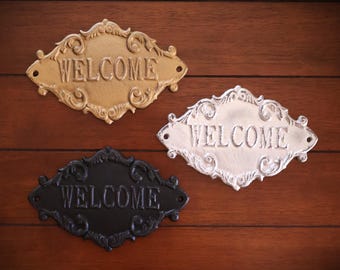 Puede incluir: Tres letreros decorativos de metal con la palabra « welcome » en oro, blanco y negro. Cada letrero tiene una forma ovalada adornada con la palabra « WELCOME » en mayúsculas. Los letreros se muestran sobre un fondo de madera.