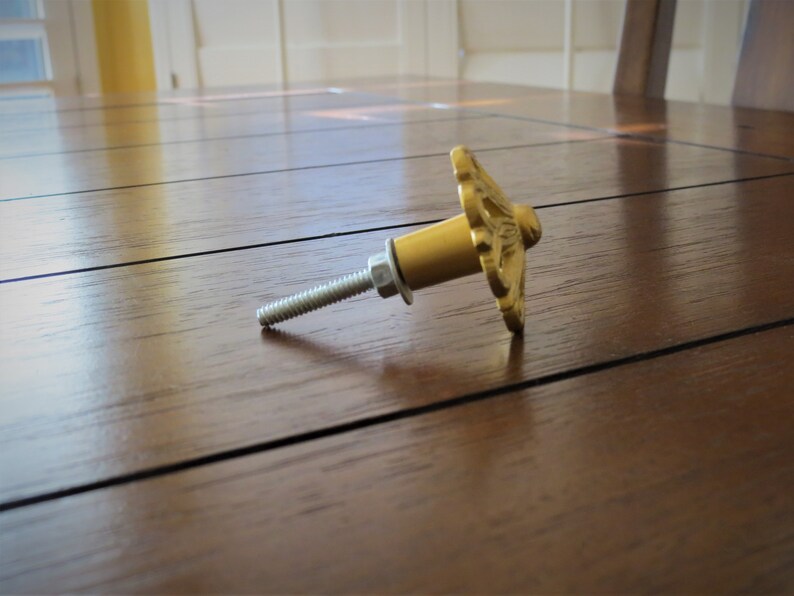 May include: Gold-toned metal cabinet knob with a decorative floral design. The knob has a screw for attaching it to a cabinet door.