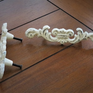 Pode incluir: Dois puxadores de gaveta de ferro fundido branco com designs ornamentados e parafusos pretos.