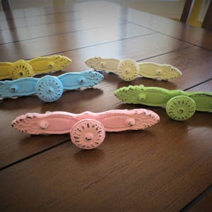 Op de afbeelding: Een set van zes pastelkleurige ladegrepen met sierlijke ontwerpen. De grepen zijn gemaakt van metaal en hebben een verweerde afwerking. De kleuren omvatten lichtblauw, geel, roze en groen.