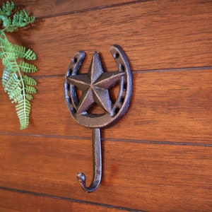 May include: A cast iron horseshoe hook with a five-pointed star in the center. The hook is painted in a dark brown color and is mounted on a wooden wall.