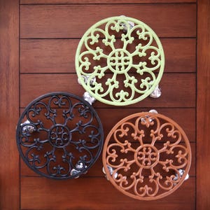 May include: Three decorative metal trivets in a circular design with ornate scrollwork. One is light green, one is black, and the third is a copper color. Each trivet has small, clear feet. The trivets are displayed on a dark wood surface.
