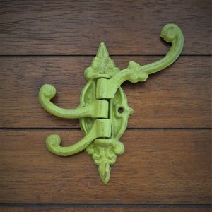 May include: A green cast iron triple hook with a decorative design. The hook is mounted on a wooden surface.