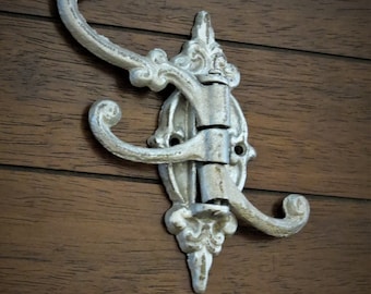 Puede incluir: Un gancho de pared decorativo de estilo antiguo con tres brazos curvos. El gancho es de metal con un acabado blanco roto envejecido. Está montado sobre una superficie de madera marrón oscura.