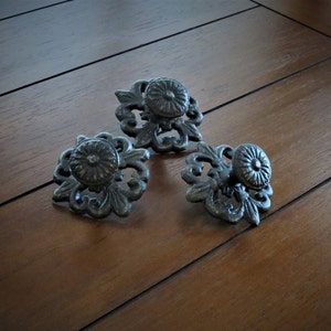 May include: Three ornate black metal drawer pulls with a floral design. Each pull has a round knob in the center.