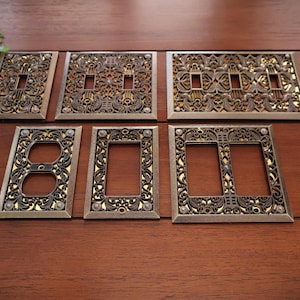 May include: A selection of decorative light switch and socket covers in a bronze finish. The covers showcase detailed floral and scroll patterns, giving a vintage appearance. Different sizes and configurations are visible.