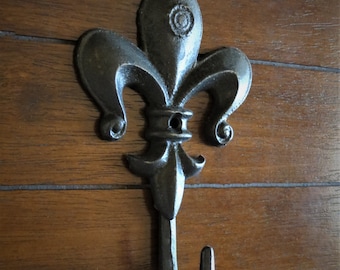 May include: Black cast iron fleur-de-lis hook with a decorative design. The hook is mounted on a wooden surface.