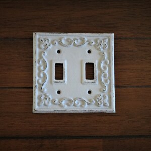 May include: White porcelain double light switch cover with an ornate design. The cover has a distressed finish and features a floral pattern.