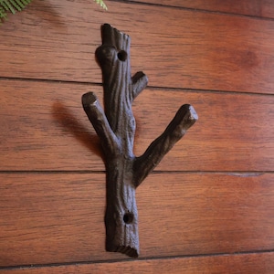 May include: A cast iron wall hook shaped like a tree branch with two hooks. The hook is dark brown and has a rustic look.