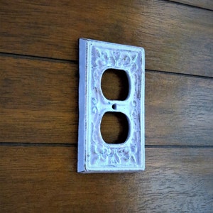 May include: White porcelain outlet cover with an ornate floral design. The cover has two standard electrical outlets.