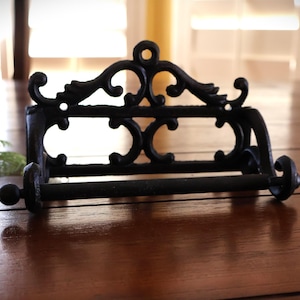 Puede incluir: Portarrollos de papel higi&eacute;nico de hierro fundido negro con un dise&ntilde;o ornamental de volutas. El soporte tiene una barra horizontal para el rollo y est&aacute; montado sobre una base rectangular. Una pieza decorativa para el ba&ntilde;o.