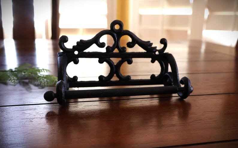 Puede incluir: Portarrollos de papel higi&eacute;nico de hierro fundido negro con un dise&ntilde;o ornamental de volutas. El soporte tiene una barra horizontal para el rollo y est&aacute; montado sobre una base rectangular. Una pieza decorativa para el ba&ntilde;o.