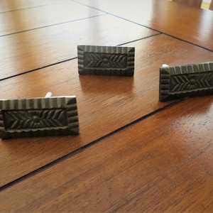 May include: Three antique-style, rectangular drawer pulls with a decorative, geometric design. The pulls are made of a dark metal with a slightly distressed finish.