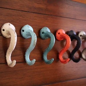 Op de afbeelding: Zeven decoratieve haken in verschillende kleuren, waaronder wit, blauw, oranje, zwart en grijs. De haken zijn S-vormig gebogen en zijn bevestigd aan een houten muur.