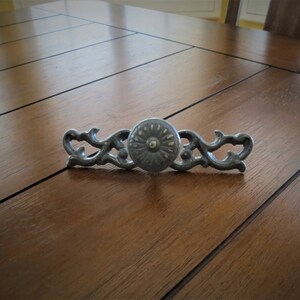 May include: A silver-toned drawer pull with a decorative floral design. The pull has a round center with a raised, circular pattern.