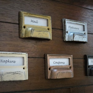 Peut inclure: Cinq tirettes de tiroir en métal vintage avec des étiquettes. Les étiquettes indiquent "Mail", "Flatware", "Napkins", "Coupons" et "Recipes".