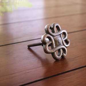 May include: A silver metal cabinet knob with a decorative, intricate design. The knob has a square shape with a rounded edge and a screw on the back.