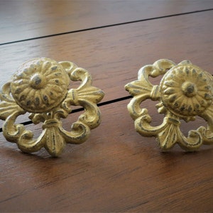 May include: Two ornate gold-toned metal drawer pulls with a floral design. Each pull has a round centre with a raised, textured pattern.