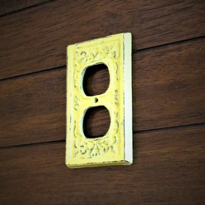 May include: A yellow painted outlet cover with an ornate design. The cover is mounted on a dark wood wall.