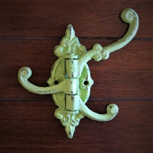 May include: A distressed, lime green metal wall hook with two curved arms and ornate detailing. The hook is mounted on a decorative backplate with a hinged design. The finish has a worn, vintage appearance.