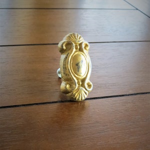 以下が含まれることがあります： 装飾的なデザインが施された金色の金属製のキャビネットノブ。ノブの中央には隆起した楕円形があり、複雑な渦巻きで囲まれています。