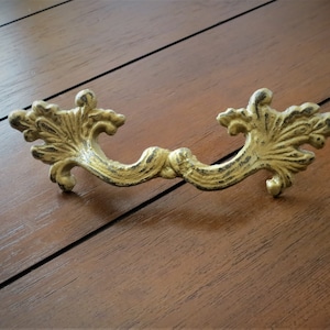 Op de afbeelding: Een gouden metalen ladegreep met een decoratief, sierlijk ontwerp. De greep heeft een gebogen vorm met twee gedetailleerde, bladvormige uiteinden.