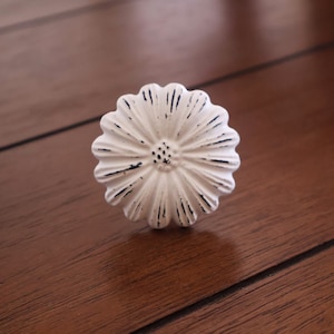 Może przedstawiać: Biały ceramiczny gałka do szuflady w kształcie kwiatu z wytartym wykończeniem. Gałka ma wypukły środek i falisty brzeg.