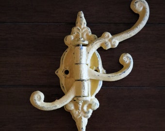 Puede incluir: Un gancho triple de hierro fundido amarillo con un diseño decorativo. El gancho está montado en una superficie de madera.