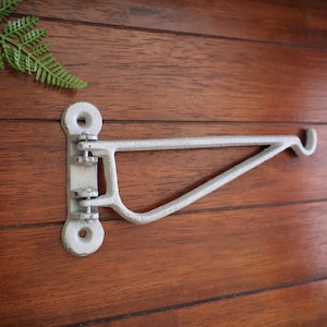 May include: White metal wall mounted shelf bracket with a hook on the end. The bracket is attached to a wooden wall.