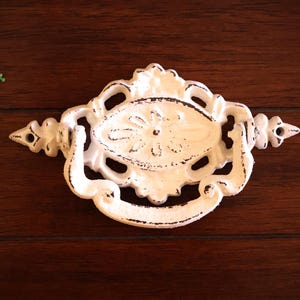 May include: White cast iron drawer pull with an ornate design. The pull has a floral pattern and a center oval.