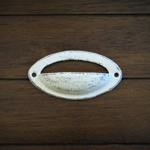 May include: White oval-shaped drawer pull with a recessed center. The pull has a distressed finish and is mounted on a dark wood surface.