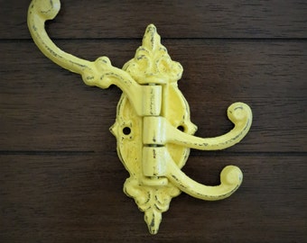 Puede incluir: Un gancho triple de metal amarillo con un diseño ornamentado. El gancho está montado en una superficie de madera.