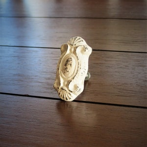Op de afbeelding: Witte keramische kastknop met een ovale vorm en ingewikkelde details. De knop heeft een verweerde afwerking, waardoor hij een vintage uitstraling heeft.