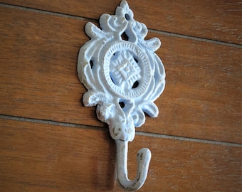 May include: A white cast iron hook with a decorative floral design. The hook is mounted on a wooden wall.