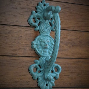 May include: A teal blue cast iron door knocker with a lion's face design. The knocker has a decorative, ornate design and is attached to a wooden door.