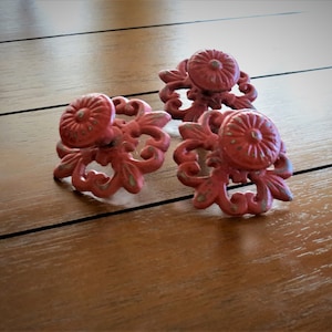 Op de afbeelding: Drie roze metalen kastknoppen met sierlijke bloemmotieven. De knoppen hebben een verweerde afwerking en zijn perfect om een vintage touch toe te voegen aan elk interieurproject.